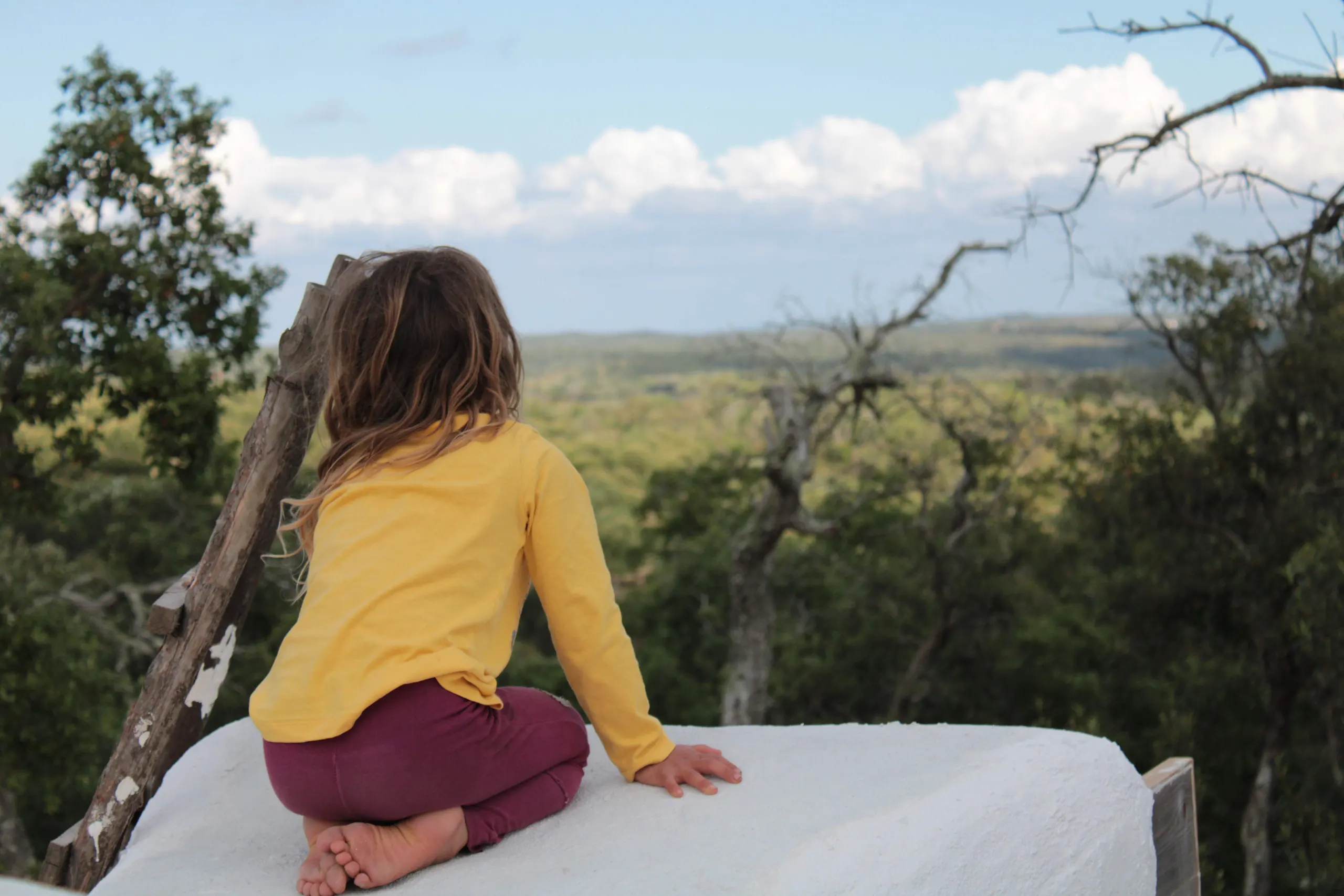 A child in a yellow top sitting barefoot on a white wall, looking out over the wooded hills