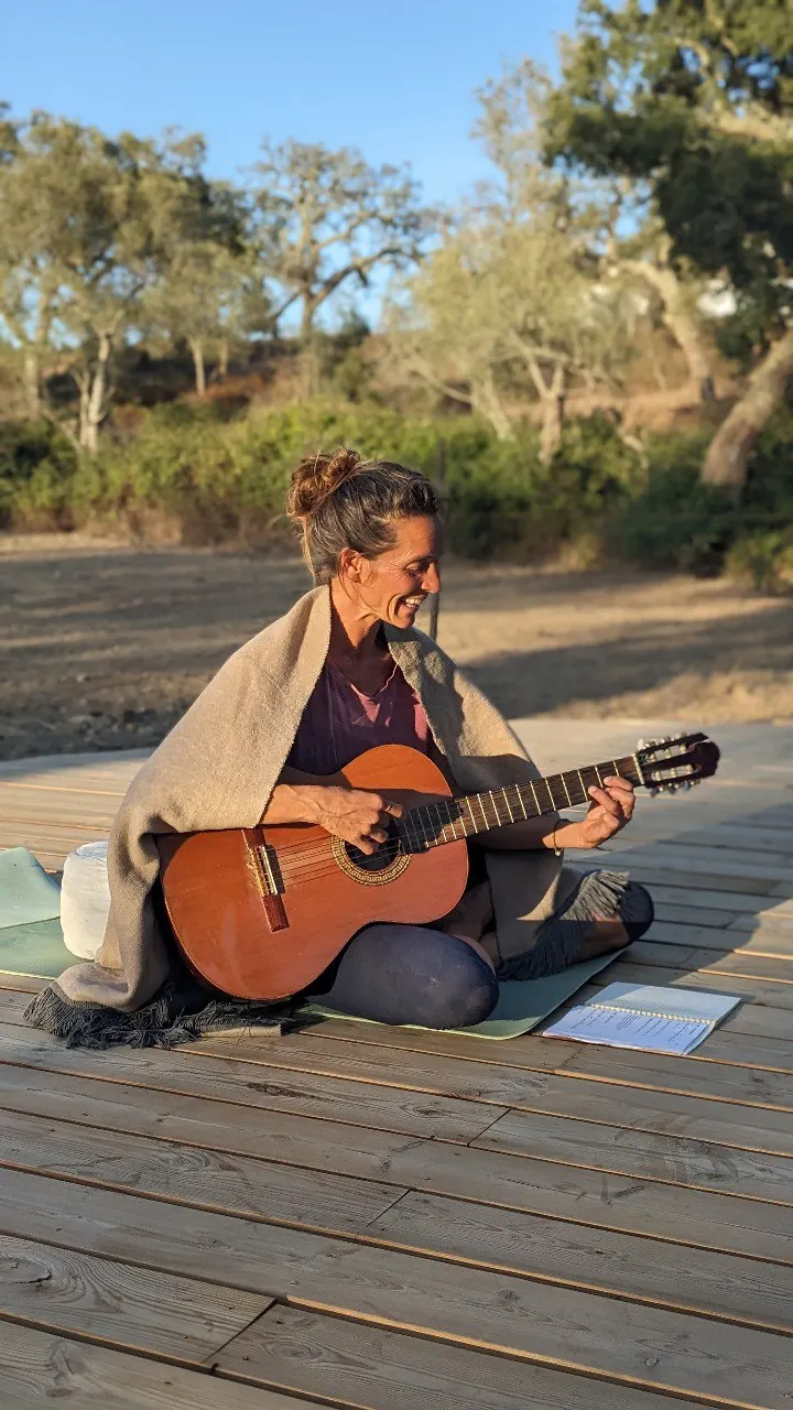 Susanne smiling and playing guitar, wrapped in a blanket on the wooden yoga deck